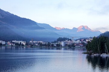 Wall Mural - Morning scenery of beautiful Lake St. Moritz, a popular resort in Engadine, Switzerland, with sunrise glow on majestic Alpine mountains in background & village buildings by lakeside in misty twilight