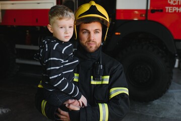 Wall Mural - Portrait of rescued little boy with firefighter man standing near fire truck. Firefighter in fire fighting operation.