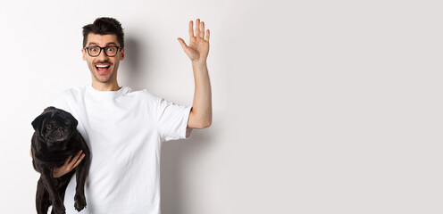 Handsome young man in glasses holding his black pug and waving hand, guy saying hello while carry dog with one arm, standing over white background