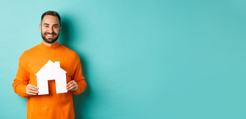 Wall Mural - Real estate concept. Happy young man searching for home rent, holding house paper maket and smiling, standing over blue background