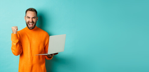 Freelance and technology concept. Lucky man winner celebrating, winning online, showing fist pump and holding laptop, standing over light blue background