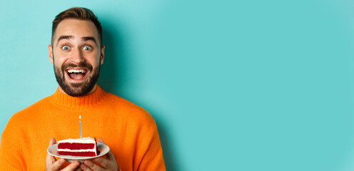 Wall Mural - Close-up of happy adult man celebrating birthday, holding bday cake with candle and making wish, standing against turquoise background