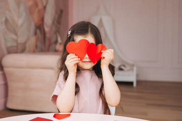 a little girl makes valentine's day cards using colored paper, scissors and pencil, sitting at a tab