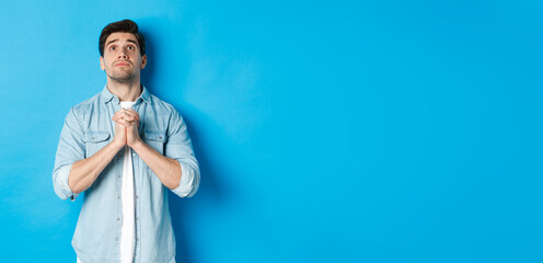 Wall Mural - Hopeful young man looking up and praying God, begging for help, standing against blue background