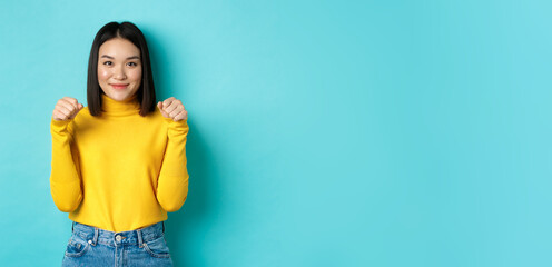 Wall Mural - Beauty and fashion concept. Beautiful and stylish asian woman in yellow pullover, holding hands raised near chest as if holding banner or logo, standing over blue background