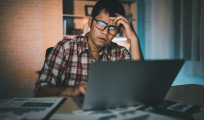 Wall Mural - Asian freelance man having stressful depression sad time working on laptop home night. Depression man sad serios working from home. Man wearing plaid shirt red is rushing work overtime for deadline.