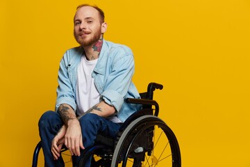 A man in a wheelchair smile and happiness, thumb up, with tattoos on his hands sits on a yellow studio background, the concept of health a person with disabilities