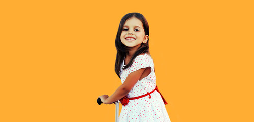 Wall Mural - Portrait of active happy little girl child skating on scooter on orange background