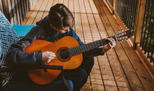 Teen Boy Playing An Acoustic Guitar On The Deck Of A Log Home.