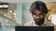 © Yuliia - Focused adult ethnic Arabian Indian businessman in glasses typing on computer working indoors. Serious male manager man executive work with laptop analyzing business digital tech data typing online