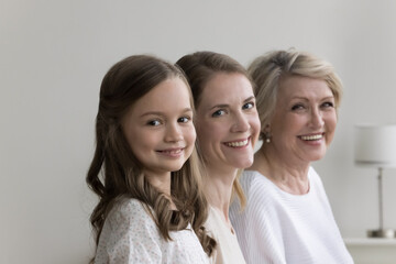 Cheerful pretty daughter kid, young mother and happy mature grandma standing in row, posing at home, looking at camera, smiling. Three female generations family portrait