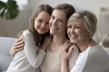 Wall Mural - Three happy girls and women of different female family generations posing together for shooting, looking away with toothy smiles, laughing, resting on home sofa, enjoying leisure, having fun