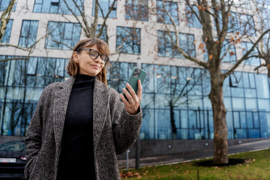 Adult beautiful businesswoman in coat and eyegkasses standing on the backdrop of a business center and using her smartphone.
