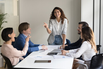 Wall Mural - Cheerful business presenter woman talking to creative team of employees, brainstorming, having fun on office meeting, pointing index fingers with elder colleague, smiling, laughing