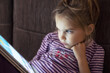 A beautiful little girl is sitting on a couch and staring at a digital tablet.