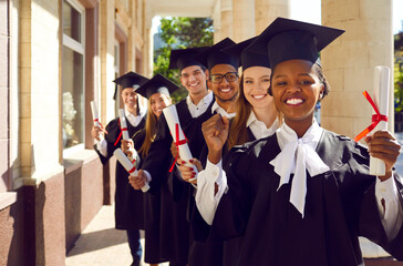 Wall Mural - Happy diverse university graduates. Six cheerful students in caps and gowns standing together near college building, holding paper scrolls, looking at camera and smiling. Graduation, success concept