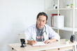 © Queenmoonlite Studio - Asian doctor sitting and writing on paper at clinic during online consultation.