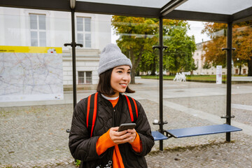 Wall Mural - Cheerful asian woman using smartphone while standing at bus station