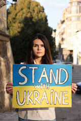 Wall Mural - Sad woman holding poster in colors of national flag and words Stand with Ukraine on city street