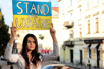 Wall Mural - Sad woman holding poster in colors of national flag and words Stand with Ukraine on city street. Space for text