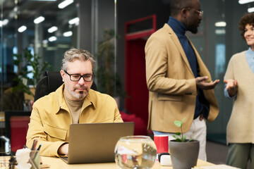 Poster - Serious mature businessman networking in front of laptop while sitting by desk against two young intercultural colleagues having discussion
