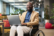 © pressmaster - Happy young black man with disability sitting in wheelchair in office and looking at camera while analyzing online data on laptop screen