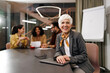 © bnenin - Smiling mature woman sitting at the office, people in the background talking.