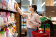 Side view of young attractive caucasian woman wearing eyeglasses holds cart and reach hand to take food from top shelf. Concept of shopping in supermarket and consumerism