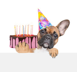 Poster - French bulldog puppy wearing party cap holding birthday cake with many candles looking above empty banner. isolated on white background