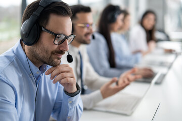 Wall Mural - Close up head shot of handsome male customer support agent with headset working in call center. Group of helpdesk operators at costumer suport service.