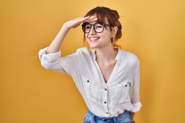 Wall Mural - Young beautiful woman wearing casual shirt over yellow background very happy and smiling looking far away with hand over head. searching concept.