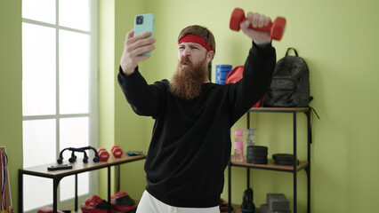 Wall Mural - Young redhead man using dumbbells training and making selfie by smartphone at sport center