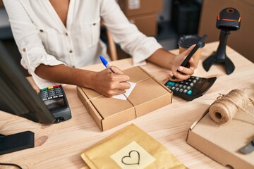 Wall Mural - Young hispanic woman ecommerce business worker using smartphone writing on package at office