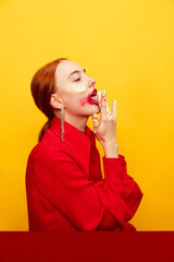 Wall Mural - Beautiful young girl in red shirt with smudged red lipstick, licking whipped cream over yellow studio background. Food pop art photography. Complementary colors. Concept of art, beauty, food.