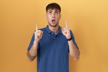 Sticker - Young hispanic man standing over yellow background amazed and surprised looking up and pointing with fingers and raised arms.