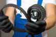 Auto mechanic in blue jumpsuit holds in his hands in black gloves roller belt tensioner of gas distribution mechanism of engine and belt. Concept of replacing belt in engine in car.