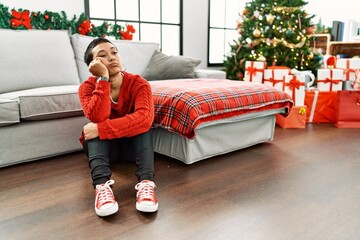 Wall Mural - Young hispanic woman unhappy sitting by christmas tree at home