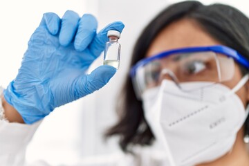 Canvas Print - Young latin woman wearing scientist uniform and medical mask holding dose vaccine at laboratory