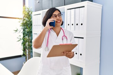 Poster - Young latin woman wearing doctor uniform talking on the smartphone at clinic