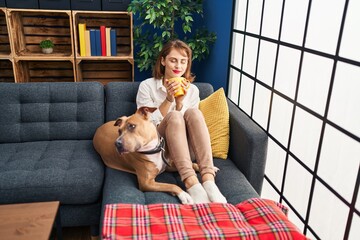 Wall Mural - Young caucasian woman drinking coffee sitting on sofa with dog at home