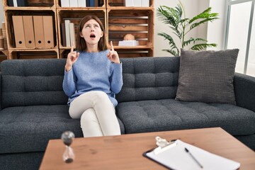 Poster - Young brunette woman at consultation office amazed and surprised looking up and pointing with fingers and raised arms.