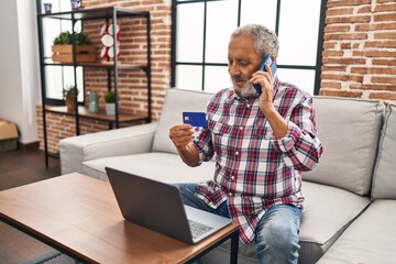 Canvas Print - Senior grey-haired man talking on smartphone holding credit card sitting on sofa at home