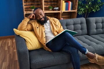 Poster - Young african american man reading book sitting on sofa at home