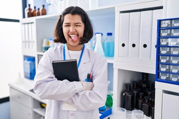 Canvas Print - Young hispanic woman working at scientist laboratory sticking tongue out happy with funny expression.