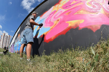 Young caucasian male graffiti artist drawing big street art painting in blue and pink tones. Stylish man in denim shorts and grey t shirt made new graffitti artwork outdoors in bright sunny day