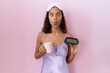 Young hispanic woman wearing nightgown holding alarm clock making fish face with mouth and squinting eyes, crazy and comical.