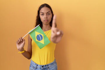 Wall Mural - Young hispanic woman holding brazil flag pointing with finger up and angry expression, showing no gesture