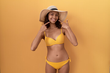 Wall Mural - Young hispanic woman wearing bikini and summer hat smiling cheerful showing and pointing with fingers teeth and mouth. dental health concept.