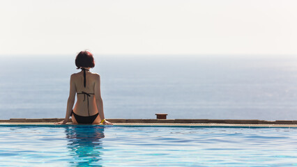 Wall Mural - A woman in a black swimsuit is sitting with her back turned on the edge of the pool.