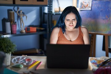 Sticker - Young modern girl with blue hair sitting at art studio with laptop at night with hand on stomach because nausea, painful disease feeling unwell. ache concept.